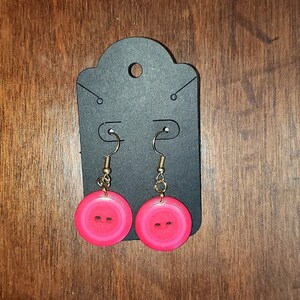 May include: A pair of pink button earrings with gold hooks. The buttons have a raised design and a shiny finish.