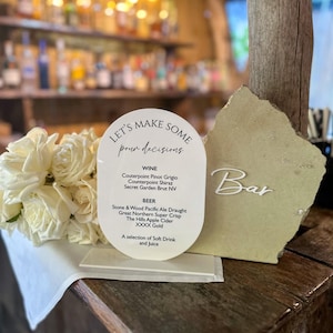 May include: An oval white acrylic menu sign on a stand with the text "LET'S MAKE SOME pour decisions" listing wine and beer options, accompanied by a bouquet of white roses and a rough-edged stone plaque with the white text "Bar" on it.