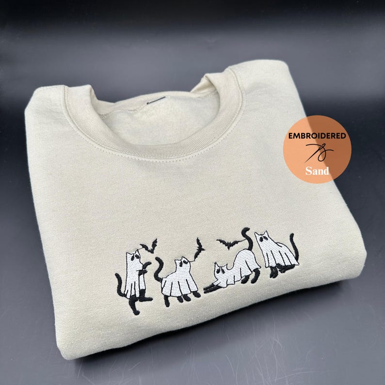 Peut inclure: Un sweat-shirt beige clair avec des chats fant&ocirc;mes et des chauves-souris brod&eacute;s en noir. Le sweat-shirt porte le texte "EMBROIDERED" et "Sand" sur le devant.