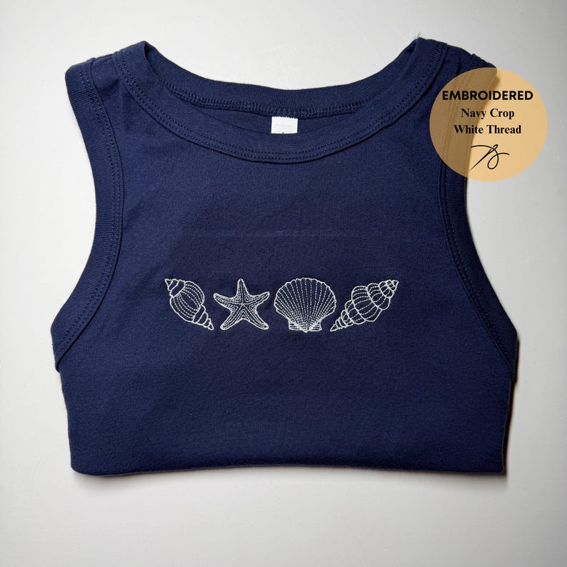 May include: Navy blue crop top with white embroidered seashells and a starfish design. The top has a round neckline and sleeveless design. The text "EMBROIDERED Navy Crop White Thread" is in a circle on the top right.