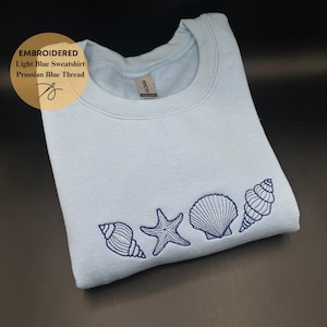 Może przedstawiać: Jasnoniebieska bluza z haftowanymi muszlami i rozgwiazdą w kolorze niebieskim pruskim. Na zdjęciu widnieje napis: "Haftowana jasnoniebieska bluza niebieska nić pruska".
