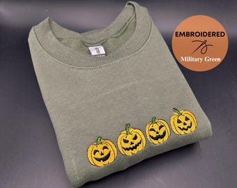 Op de afbeelding: Een groen sweatshirt met een geborduurd ontwerp van vier glimlachende pompoenen. Het sweatshirt is gelabeld met "Embroidered" en "Military Green".