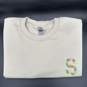 Può includere: Una felpa beige con una lettera "S" ricamata con fiori sul lato destro.