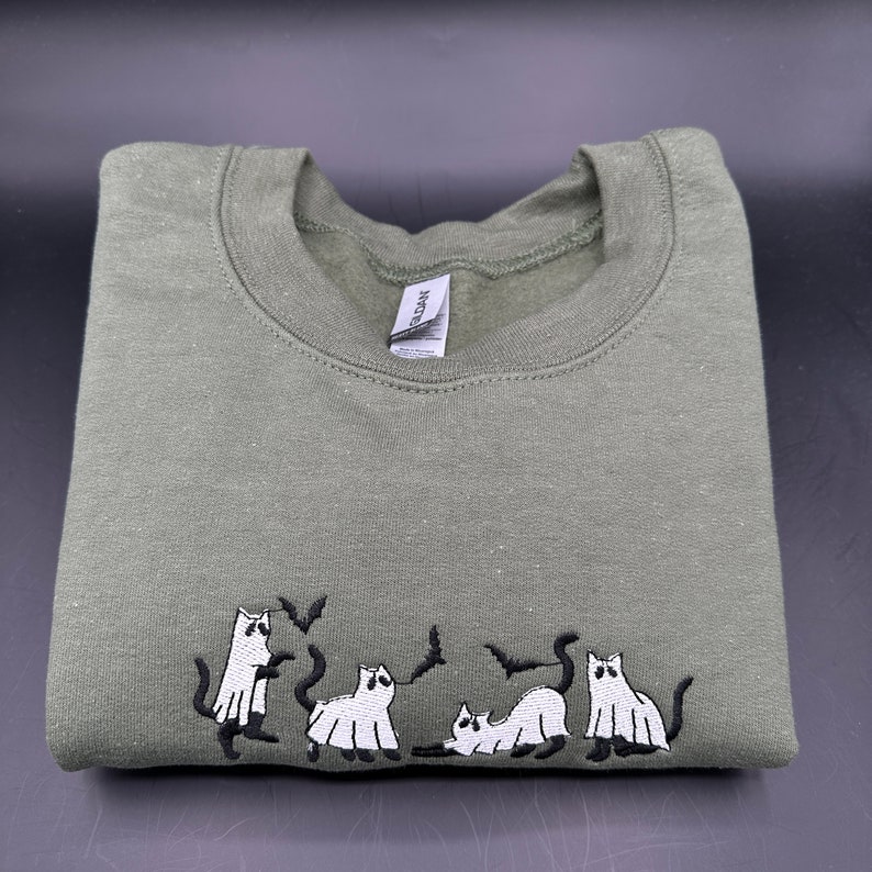 Peut inclure: Un sweat-shirt vert avec un motif brod&eacute; de quatre chats blancs habill&eacute;s en fant&ocirc;mes avec des chauves-souris qui volent autour d'eux.