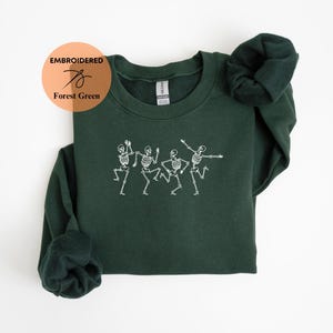 May include: A forest green sweatshirt with an embroidered design of four skeletons dancing. The sweatshirt is a crew neck style with long sleeves.