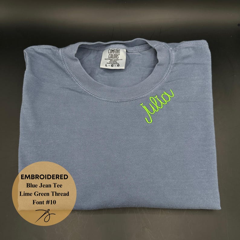 May include: A folded blue jean tee shirt with the name "Julia" embroidered in lime green thread. The shirt has a crew neck and a tag that reads "COMFORT COLORS". The text "EMBROIDERED Blue Jean Tee Lime Green Thread Font #10" is also visible.