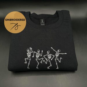 May include: Black t-shirt with white embroidered skeletons dancing. The skeletons are in a line and are all facing the same direction. The t-shirt is folded in half and is lying on a flat surface.