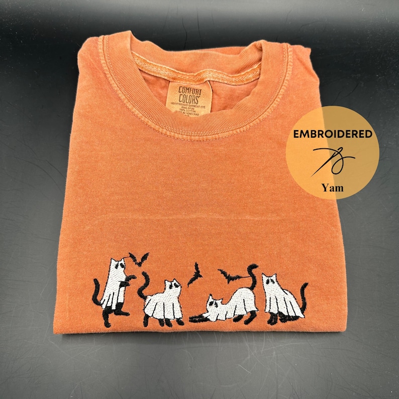 Peut inclure: T-shirt orange avec des chats fant&ocirc;mes blancs et des chauves-souris brod&eacute;s. Le t-shirt est fabriqu&eacute; par Comfort Colors et porte l'inscription "Embroidered" avec un motif d'aiguille et de fil et "Yam" en dessous.