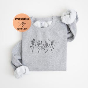 May include: A gray sweatshirt with an embroidered design of four skeletons dancing. The sweatshirt is folded and the tag says "Embroidered Sport Gray".