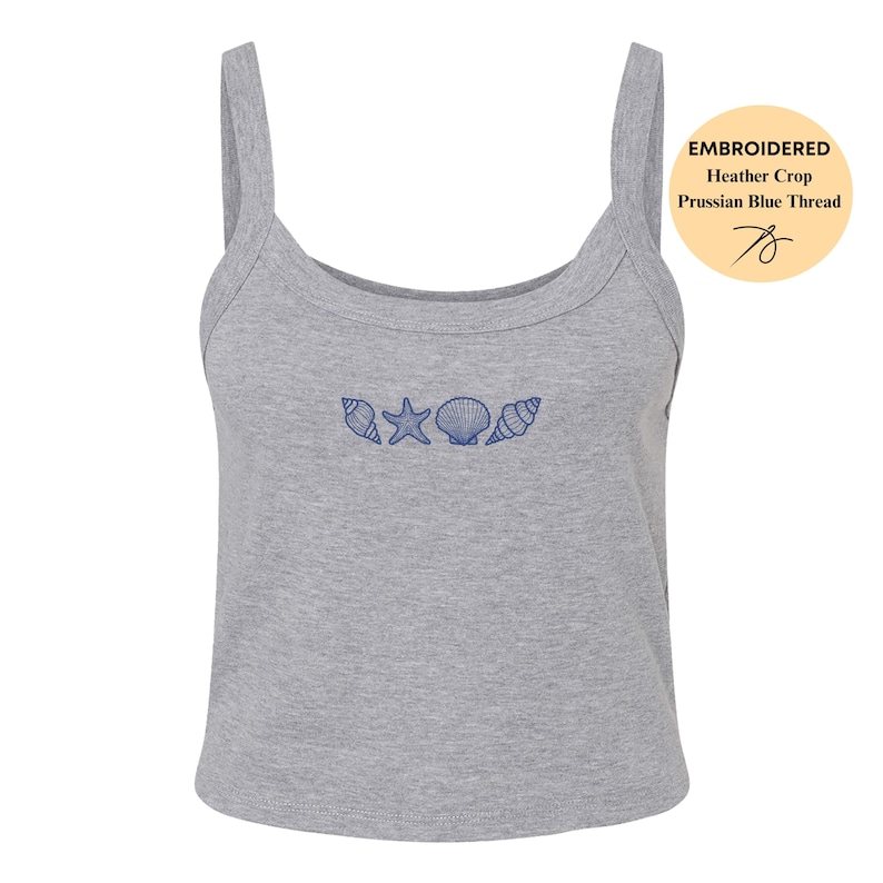 May include: A heather gray crop top with a scoop neckline and thin straps. The top is embroidered with Prussian blue thread, featuring a seashell, starfish, and two other shell designs. The image includes a circular tag with the words "EMBROIDERED Heather Crop Prussian Blue Thread".