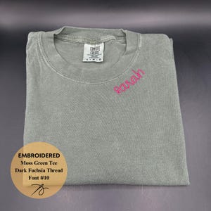 May include: A folded moss green t-shirt with the name "Sarah" embroidered in dark fuchsia thread. The shirt has a crew neck and a tag that reads "Comfort Colors". The image also includes a graphic with the text "Embroidered Moss Green Tee Dark Fuchsia Thread Font #10".