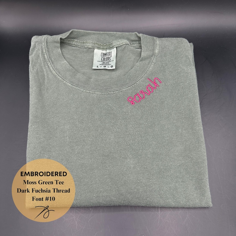 May include: A folded moss green t-shirt with the name "Sarah" embroidered in dark fuchsia thread. The shirt has a crew neck and a tag that reads "Comfort Colors". The image also includes a graphic with the text "Embroidered Moss Green Tee Dark Fuchsia Thread Font #10".