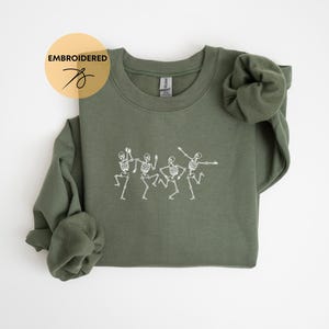 May include: A green sweatshirt with an embroidered design of four skeletons dancing. The sweatshirt has a crew neck and long sleeves.