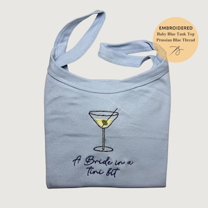 Op de afbeelding: Babyblauwe tanktop met geborduurd martini glasontwerp en de tekst "A Bride in a Tini Bit". De top heeft bandjes en een ronde halslijn. De afbeelding bevat ook een cirkelvormige afbeelding met de tekst "Embroidered Baby Blue Tank Top Prussian Blue Thread".