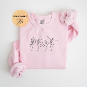 May include: Pink crewneck sweatshirt with an embroidered design of four skeletons dancing. The sweatshirt has a relaxed fit and is made of a soft, comfortable fabric.