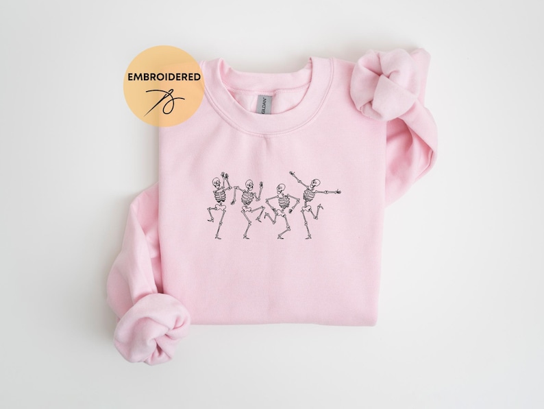 May include: Pink crewneck sweatshirt with an embroidered design of four skeletons dancing. The sweatshirt has a relaxed fit and is made of a soft, comfortable fabric.