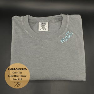 May include: A gray Comfort Colors t-shirt with the name "matt" embroidered in light blue thread. The shirt is folded neatly, with a tag visible at the neckline. The text "EMBROIDERED Gray Tee Light Blue Thread Font #10" is in a gold circle.