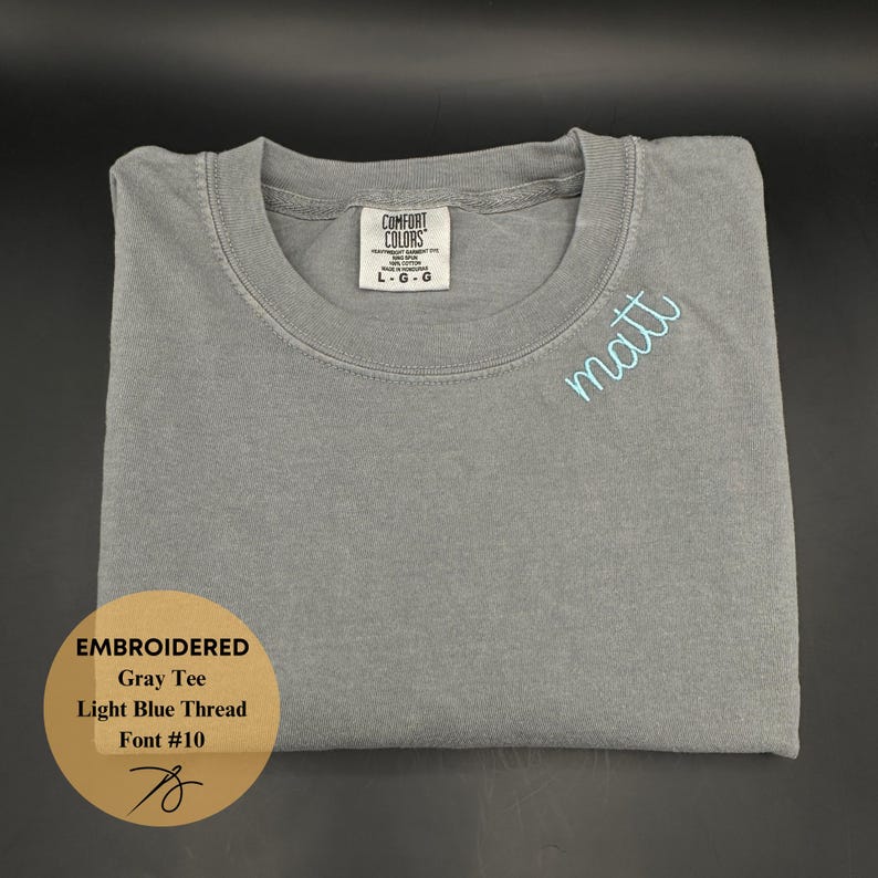 May include: A gray Comfort Colors t-shirt with the name "matt" embroidered in light blue thread. The shirt is folded neatly, with a tag visible at the neckline. The text "EMBROIDERED Gray Tee Light Blue Thread Font #10" is in a gold circle.