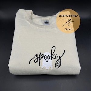 May include: A beige sweatshirt with the word "spooky" embroidered in black cursive lettering. A white ghost with a black eye is embroidered next to the word. The text "Embroidered Sand" is in a circle in the top right corner.