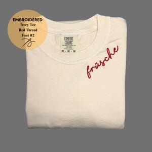 May include: Ivory tee shirt with the word "fr&auml;sche" embroidered in red thread. The shirt has a crew neck and is folded. The text "EMBROIDERED Ivory Tee Red Thread Font #2" is in a circle on the upper left.