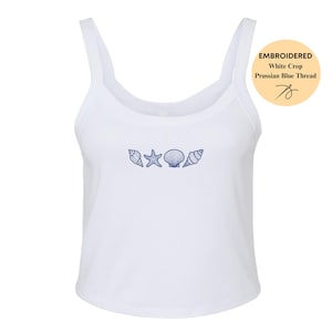 May include: White crop top with a scoop neck and thin straps. The top features an embroidered design of seashells and a starfish in Prussian blue thread. The image includes a circular graphic that reads "Embroidered White Crop Prussian Blue Thread".