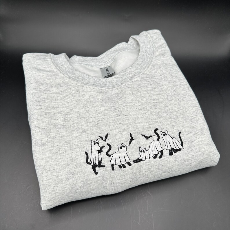 Peut inclure: Un sweat-shirt gris avec un motif brod&eacute; noir de cinq chats habill&eacute;s en fant&ocirc;mes. Les chats portent des costumes de fant&ocirc;mes avec des chauves-souris qui volent autour d'eux.
