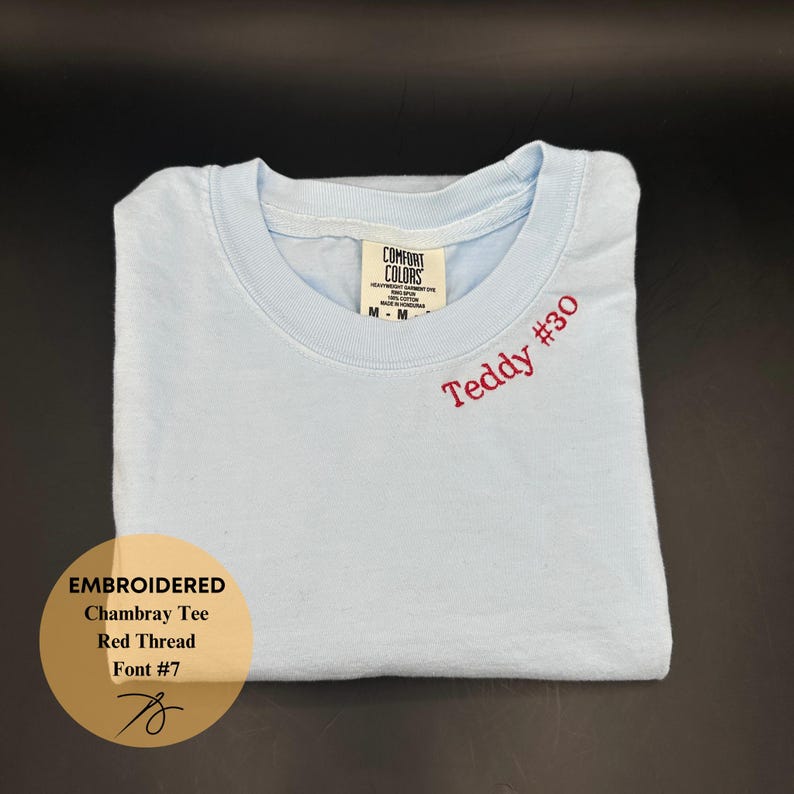 May include: A light blue chambray tee shirt with the embroidered text "Teddy #30" in red thread. The shirt is folded and has a tag that says "Comfort Colors". A graphic on the bottom left says "Embroidered Chambray Tee Red Thread Font #7".