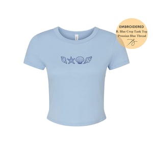 May include: A light blue crop top with embroidered seashells and a starfish design. The shirt has short sleeves and a crew neck. The text on the tag says "EMBROIDERED B. Blue Crop Tank Top Prussian Blue Thread".