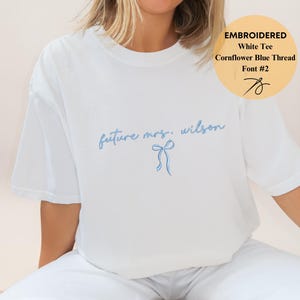 May include: White tee shirt with embroidered text "future mrs. wilson" in cornflower blue thread. A blue bow is embroidered below the text. The shirt also has a circular graphic with the words "EMBROIDERED White Tee Cornflower Blue Thread Font #2".