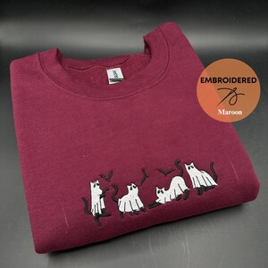 Peut inclure: Sweat-shirt bordeaux avec des chats fant&ocirc;mes blancs et des chauves-souris brod&eacute;s. Le texte "EMBROIDERED" est dans un cercle avec un graphique d'aiguille et de fil et le mot "Maroon" en dessous.