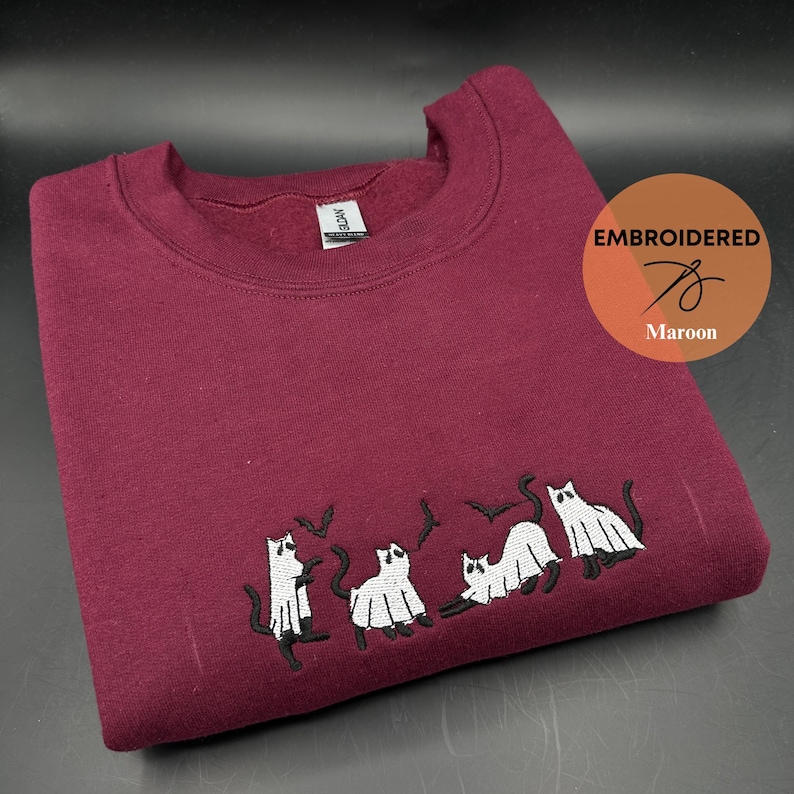 Peut inclure: Sweat-shirt bordeaux avec des chats fant&ocirc;mes blancs et des chauves-souris brod&eacute;s. Le texte "EMBROIDERED" est dans un cercle avec un graphique d'aiguille et de fil et le mot "Maroon" en dessous.