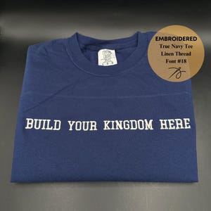 May include: A navy blue t-shirt with white embroidery that reads "BUILD YOUR KINGDOM HERE". The shirt is made of linen thread and has a tag that says "Cotton Colors".