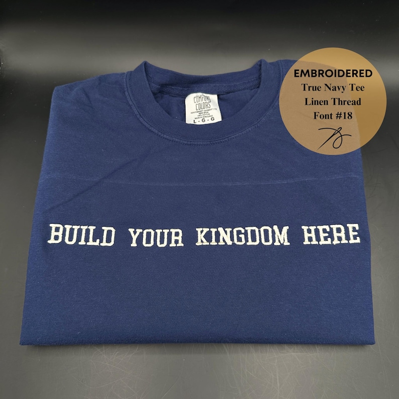 May include: A navy blue t-shirt with white embroidery that reads "BUILD YOUR KINGDOM HERE". The shirt is made of linen thread and has a tag that says "Cotton Colors".