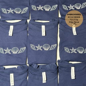 May include: Navy blue crop tops with white embroidered seashell and starfish designs. The shirts are arranged in a grid, showcasing the beach-themed embroidery. The tags read "BELLA+CANVAS". The text "EMBROIDERED BULK ORDER Navy Crop White Thread" is also visible.