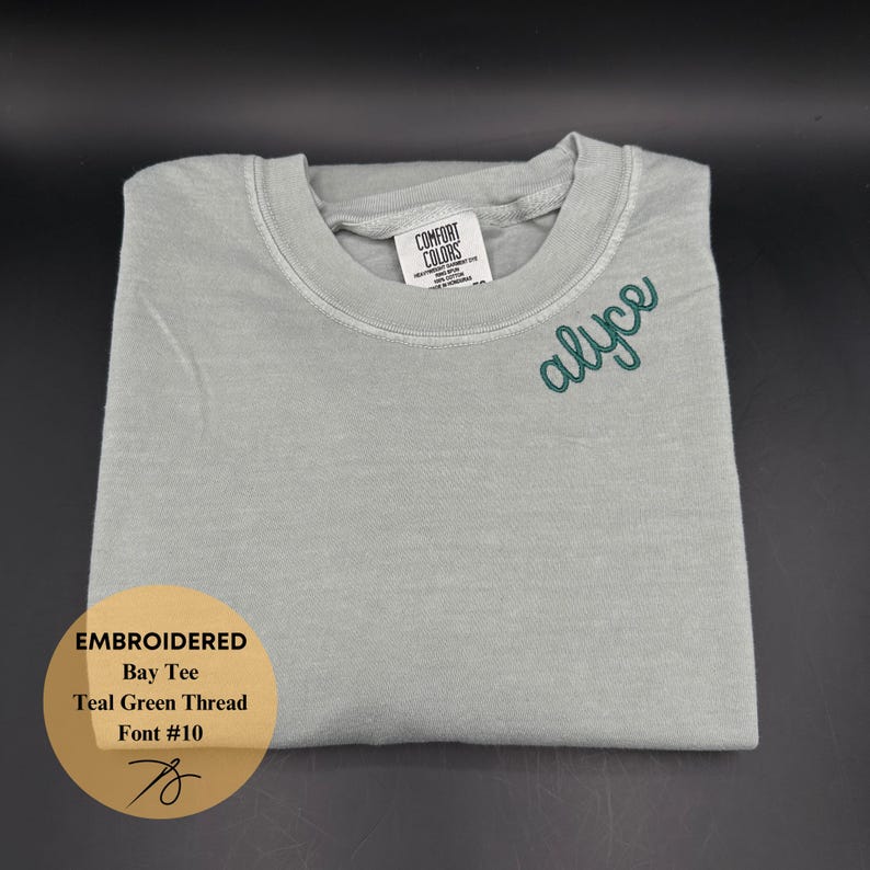 May include: A folded, light gray Bay Tee shirt with the name "alyce" embroidered in teal green thread. The shirt has a Comfort Colors tag and a circular sticker with details about the embroidery.