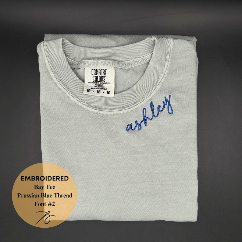 May include: A light gray Comfort Colors t-shirt with the name "ashley" embroidered in Prussian blue thread. The shirt is folded neatly and features a crew neck. The image also includes text that reads "Embroidered Bay Tee, Prussian Blue Thread, Font #2".