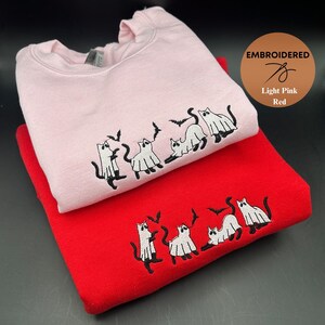 Peut inclure: Deux sweats &agrave; capuche avec des images brod&eacute;es de chats habill&eacute;s en fant&ocirc;mes. Le sweat-shirt du haut est rose clair et le sweat-shirt du bas est rouge. Le texte "EMBROIDERED" est visible dans le coin sup&eacute;rieur droit de l'image.