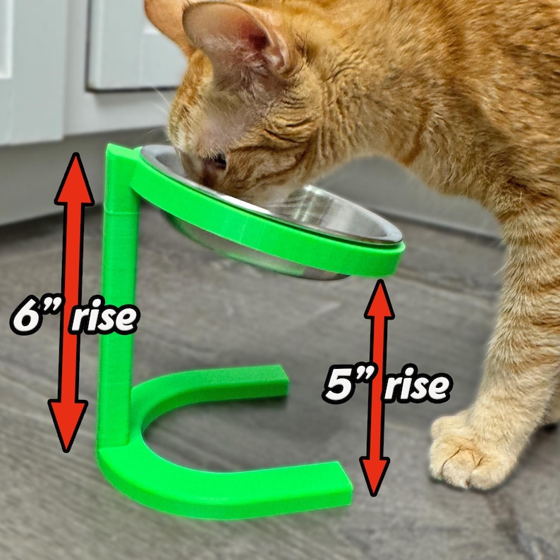 May include: A green elevated pet bowl with a stainless steel bowl, designed for cats. The stand has a 15 cm and 13 cm rise. An orange tabby cat is eating from the bowl.