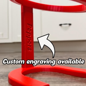 May include: A vibrant red, curved stand with the word "Peppers" engraved on its side. The text "Custom engraving available" is displayed with a white arrow pointing towards the stand. The stand is likely for display purposes.