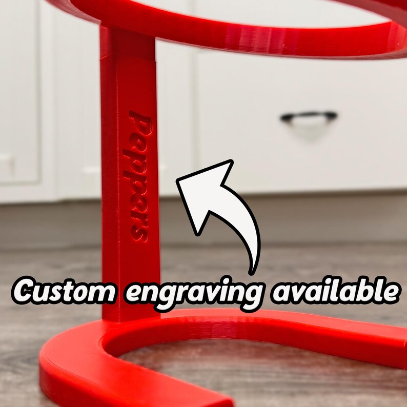 May include: A vibrant red, curved stand with the word "Peppers" engraved on its side. The text "Custom engraving available" is displayed with a white arrow pointing towards the stand. The stand is likely for display purposes.