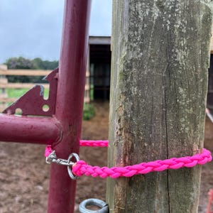 May include: A bright neon pink braided rope gate latch featuring a silver metal swivel snap hook. The rope is wrapped around a weathered wooden post and secured to a maroon metal gate frame, creating a functional closure for an outdoor enclosure.