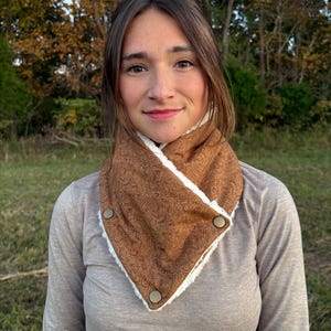 Puede incluir: Un calentador de cuello marrón y blanco con un estampado de cachemira y tres cierres de botón. El calentador de cuello es reversible con un forro de piel sintética blanca y suave. La persona de la imagen lleva una camisa de manga larga marrón claro.