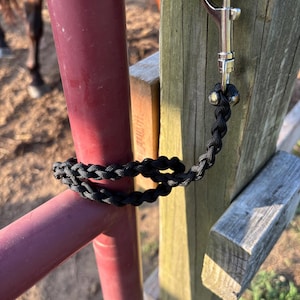 Op de afbeelding: Een zwart gevlochten touw met aan één uiteinde een zilverkleurige metalen musketonhaak. Het touw is om een rode metalen stang geslagen en aan een houten paal bevestigd, wat een duurzame constructie met een strak en uniform vlechtpatroon toont.