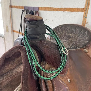 May include: A close-up of a brown leather saddle with a green and black braided rope wrapped around the horn. The saddle is sitting on a brown blanket.