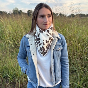 May include: A denim jacket with a cream-colored sherpa lining and a white shirt. A cow print bandana is worn around the neck. The background is a field of tall grass under a blue sky.