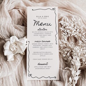 May include: A wedding menu card with a black border and elegant script. The menu includes a starter, main course, and dessert. The card is on a beige cloth with a white flower and dried floral arrangement.