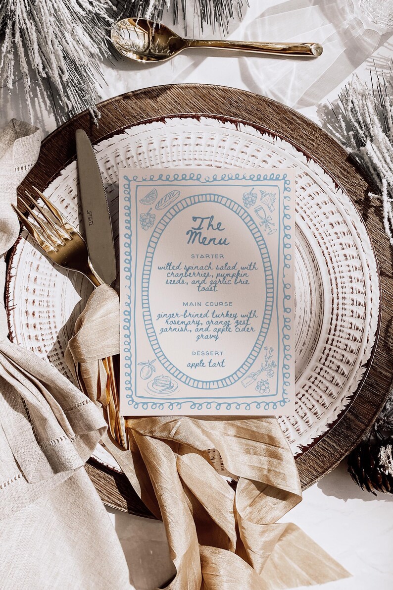 May include: A white menu card with blue and black lettering on a wooden plate with a gold napkin. The menu card has a blue border and lists the starter, main course, and dessert. The starter is wilted spinach salad with cranberries, pumpkin seeds, and garlic brie toast. The main course is ginger-brined turkey with rosemary, orange zest garnish, and apple cider gravy. The dessert is apple tart.