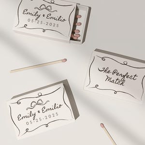 May include: Two white matchboxes with black calligraphy. One box says "Emily & Emilio 05.25.2025" with a black bow. The other box says "The Perfect Match".