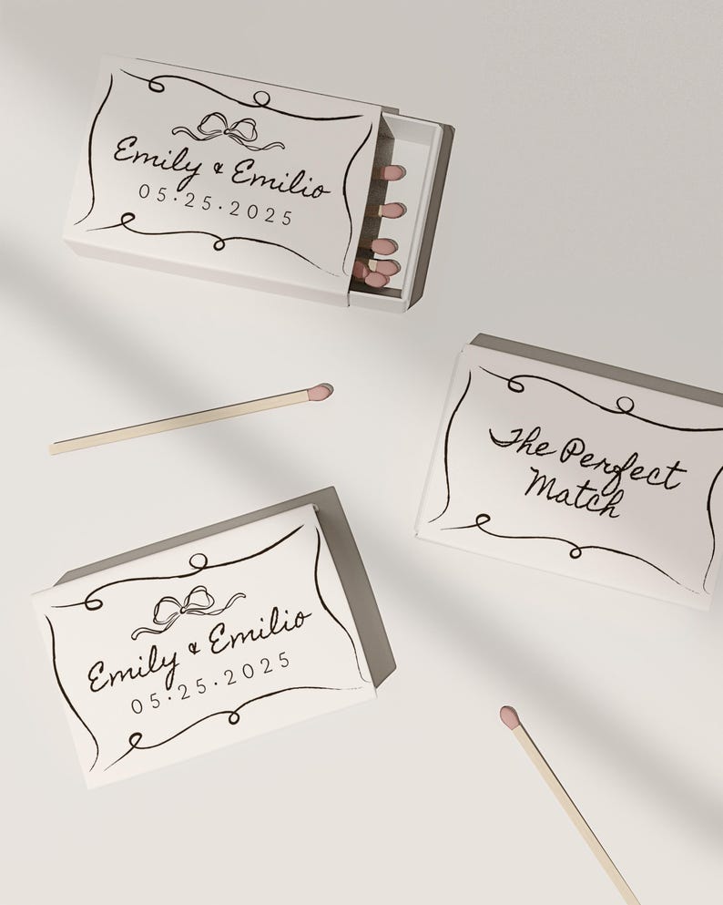 May include: Two white matchboxes with black calligraphy. One box says "Emily & Emilio 05.25.2025" with a black bow. The other box says "The Perfect Match".