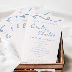 May include: A white wedding ceremony program with blue text. The program lists the order of ceremony for Emily & Emilio's wedding on May 25, 2025, at Southland.